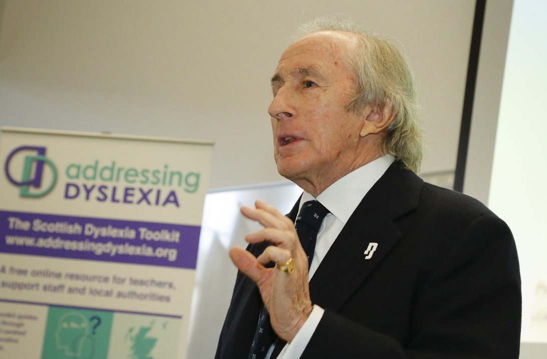En person i formell klädsel talar vid en konferens om dyslexi, med en banderoll för 'Addressing Dyslexia' i bakgrunden.