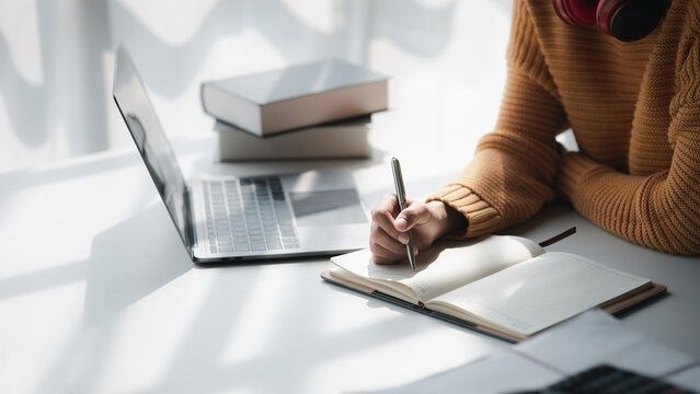 Student antecknar i en öppen skrivbok vid laptop och kursböcker